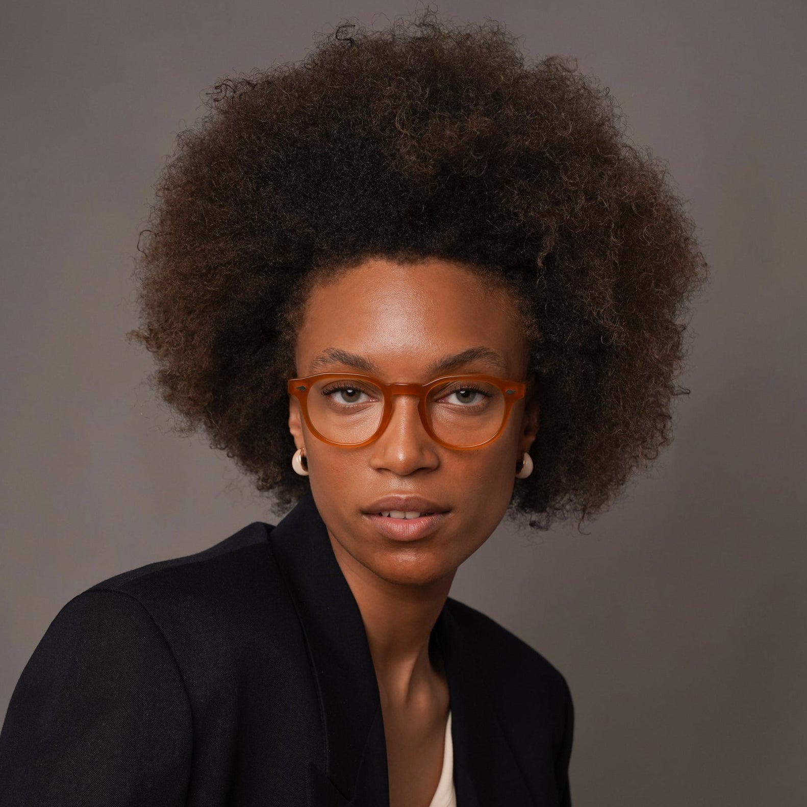 A woman with an afro hairstyle wearing orange-brown rectangular eyeglasses, a black blazer, and a cream blouse, posed against a solid dark gray studio background.