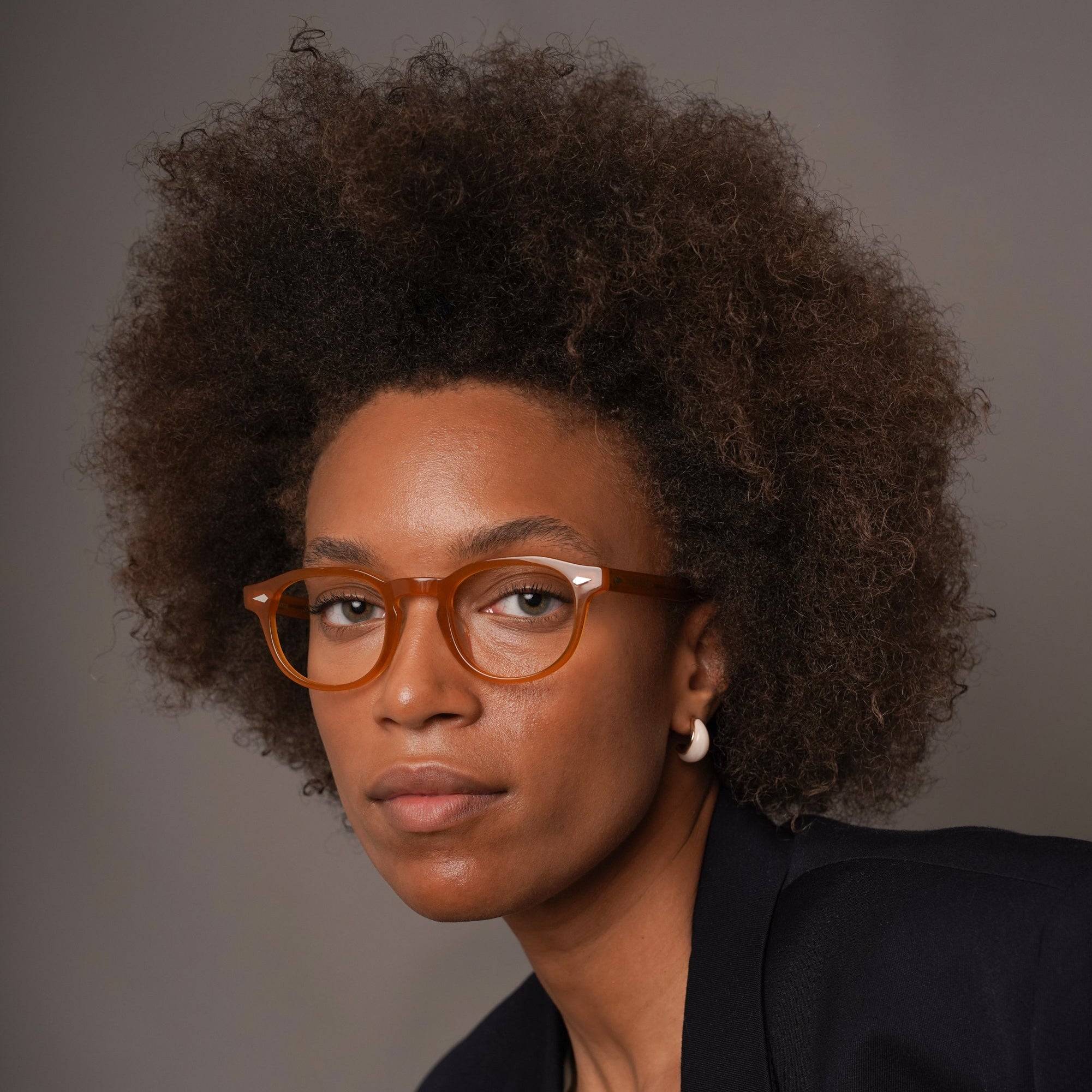A woman with an afro hairstyle wearing yellow-amber round eyeglasses, a dark navy blazer, and a cream top, photographed against a solid gray studio background.