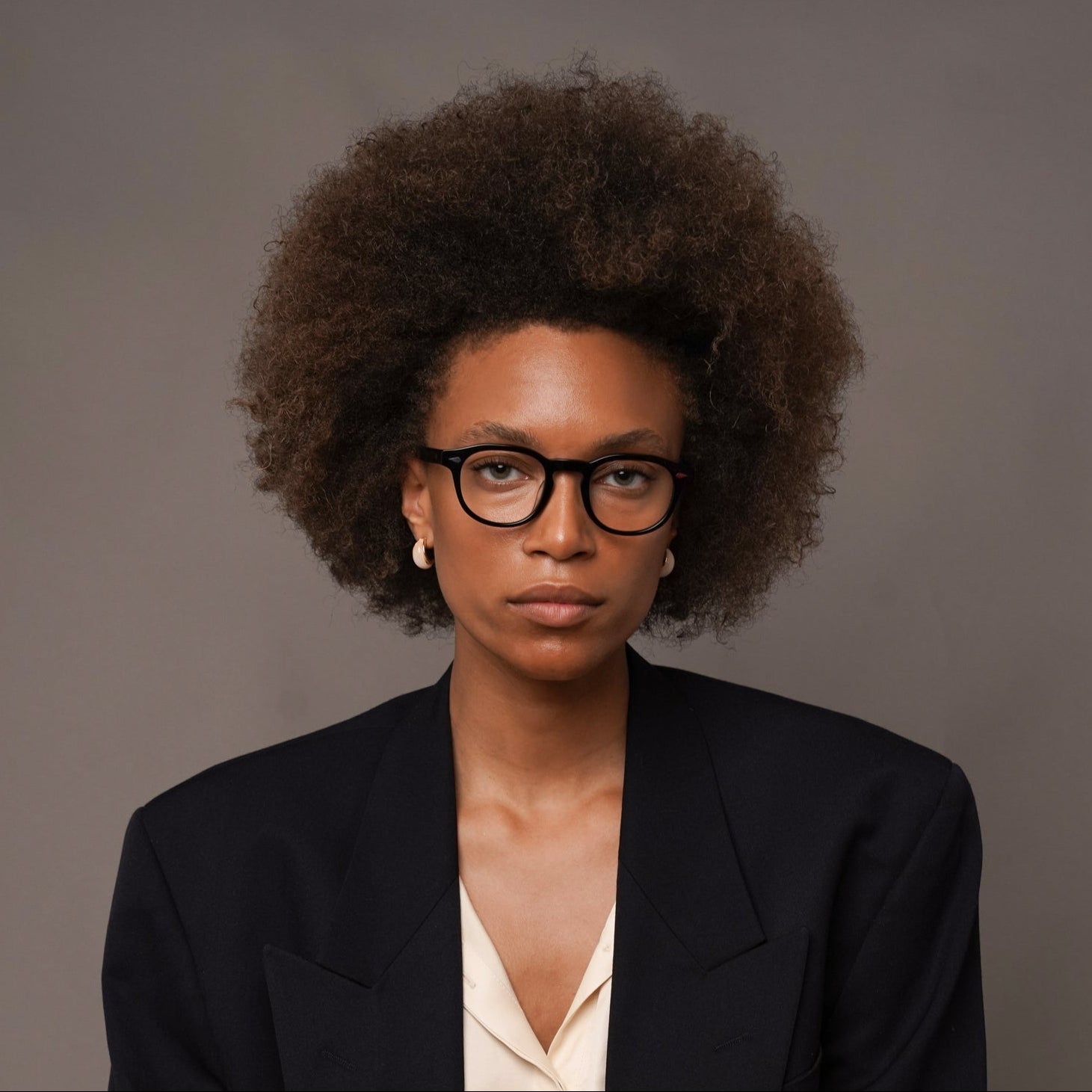 A woman with an afro hairstyle and black-rimmed glasses sits cross-legged against a neutral gray studio background. She is wearing a dark navy tailored blazer and trousers over a cream satin blouse.