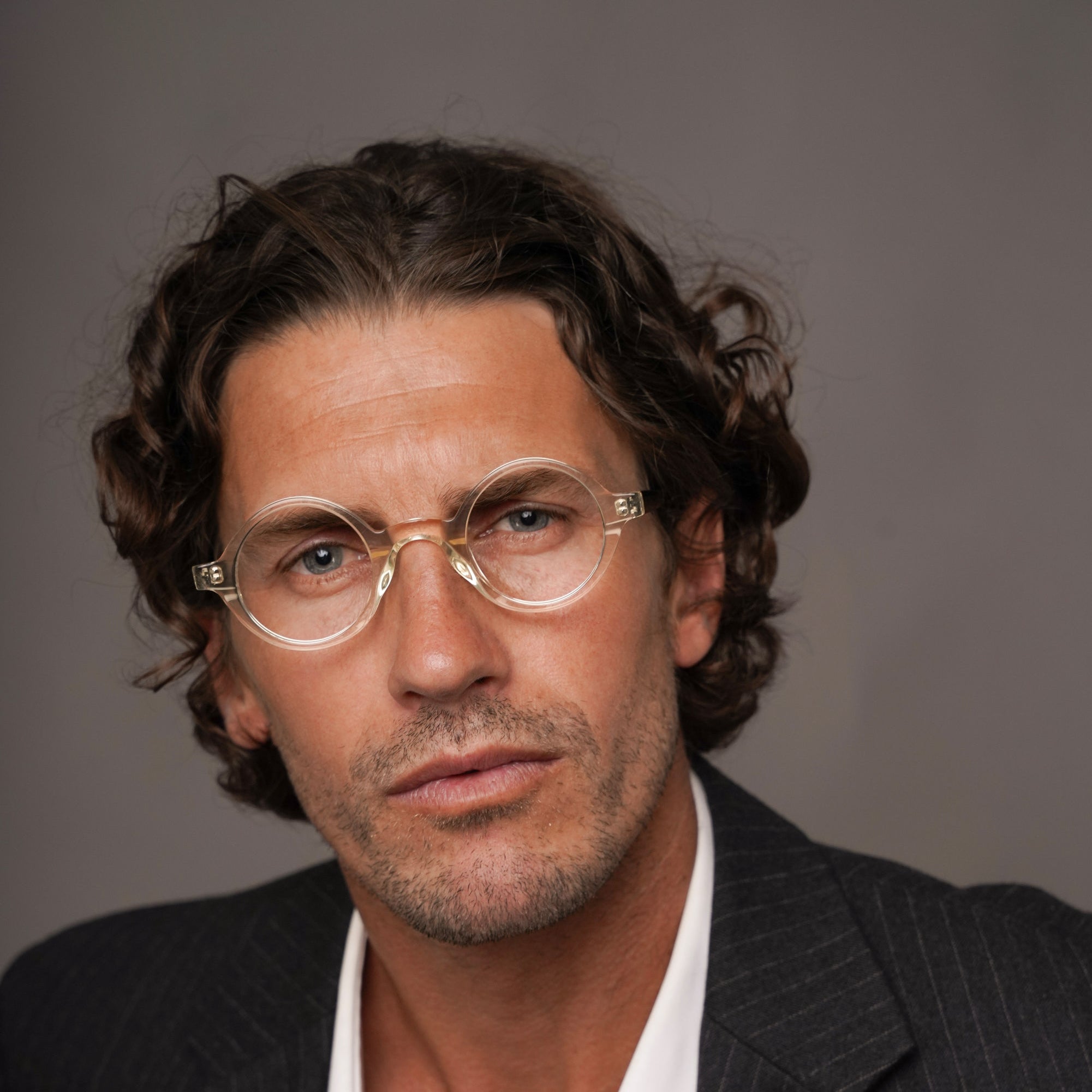 A close-up portrait of a man with curly brown hair and glasses, wearing a dark pinstripe blazer over an open white shirt, against a plain gray studio background.