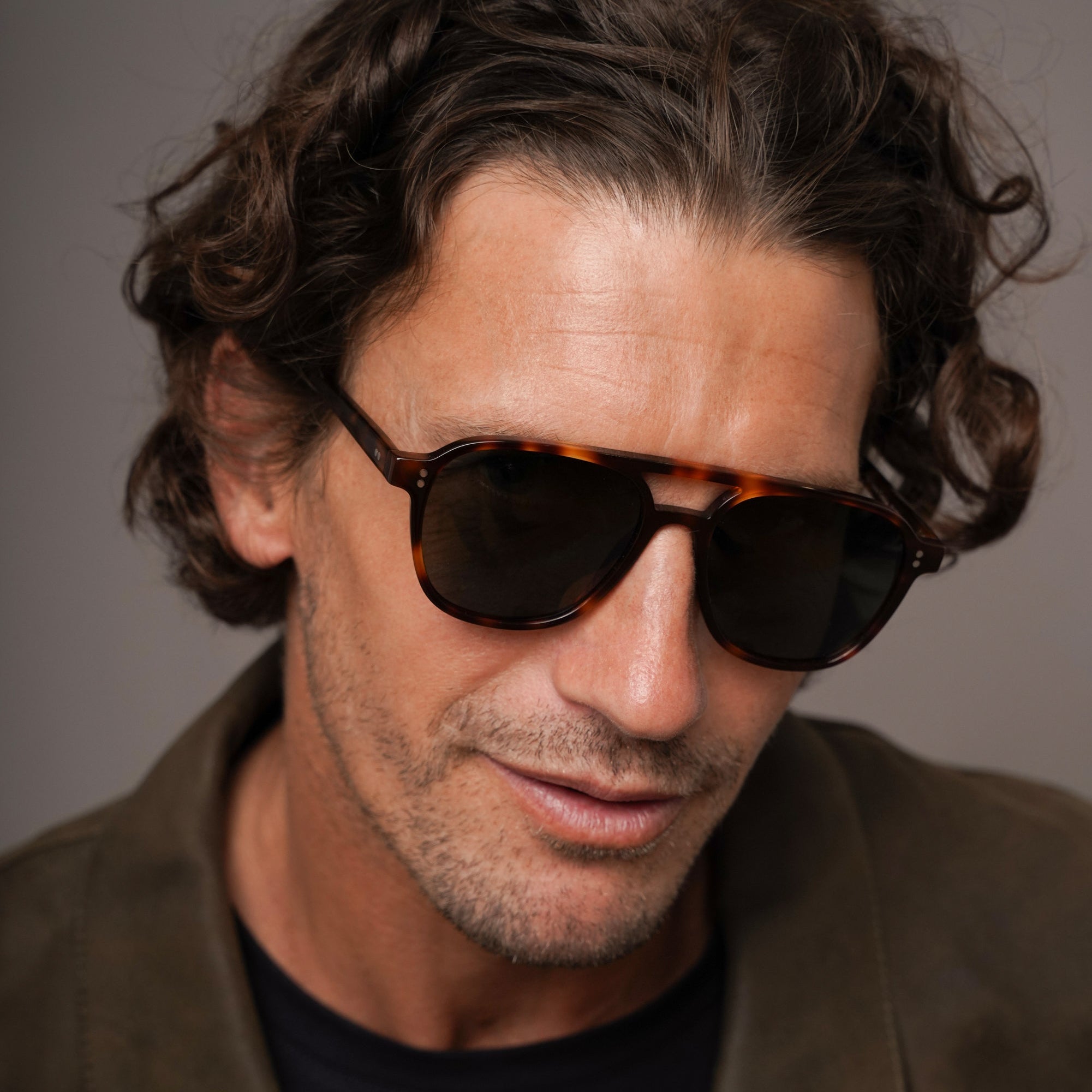 A close-up portrait of a man with curly brown hair wearing tortoiseshell sunglasses with dark lenses and a brown jacket over a black shirt, set against a plain gray background.