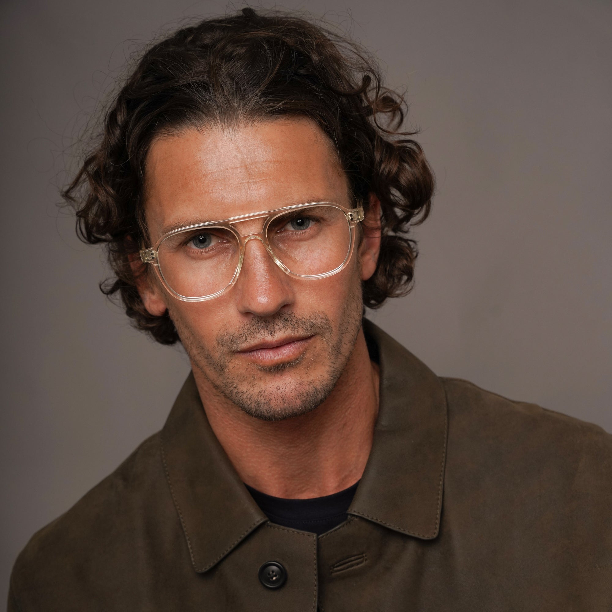 A man with curly brown hair and glasses wearing a brown suede jacket over a black shirt, photographed against a neutral gray studio background.