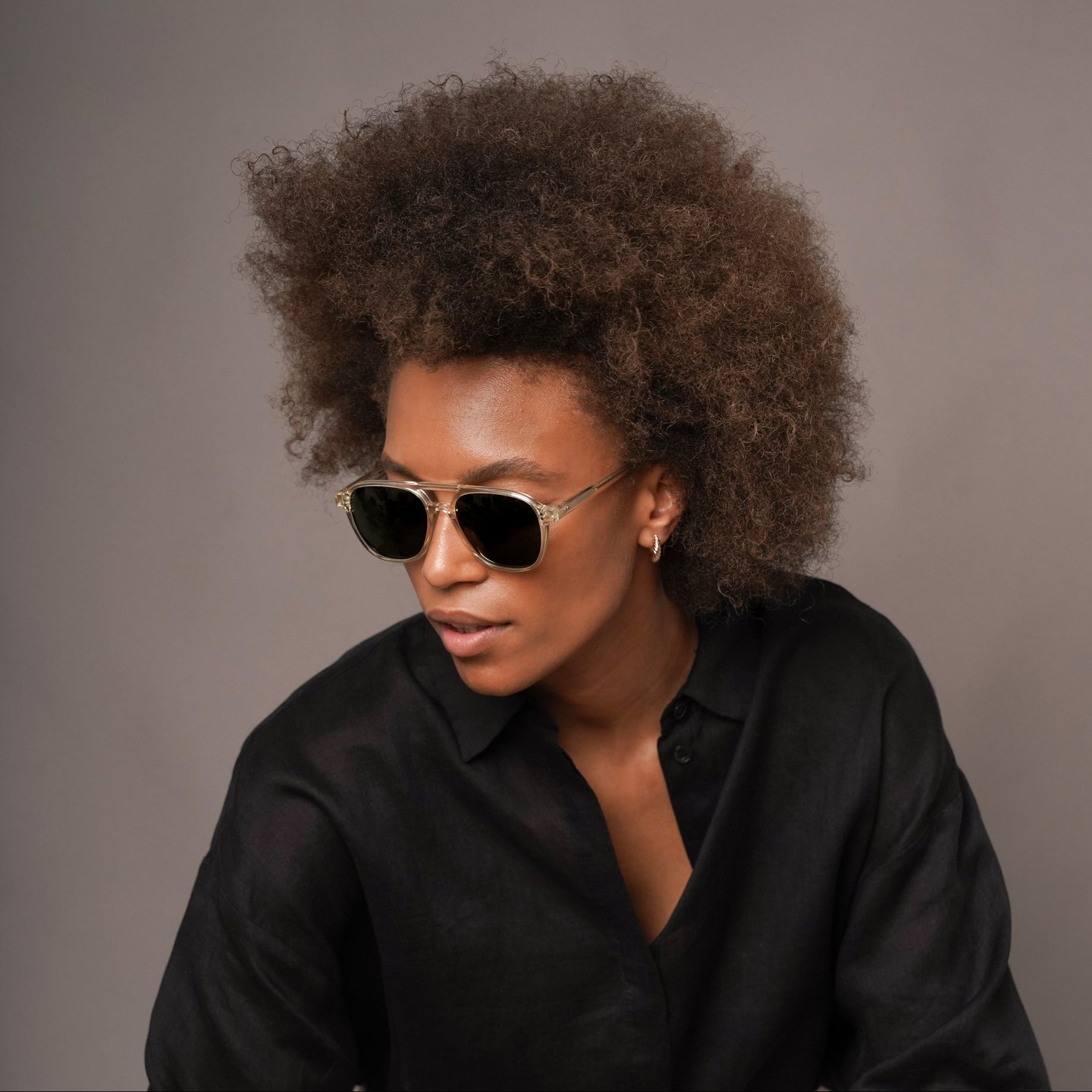 A woman with an afro hairstyle wearing clear-framed aviator sunglasses, a black button-up shirt, and off-white trousers, seated against a solid gray studio background.