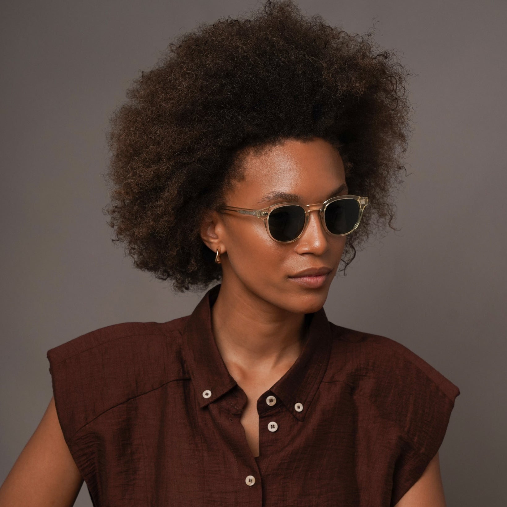 A woman with an afro hairstyle wearing round sunglasses, a dark brown sleeveless button-up shirt, and cream-colored pants, posing against a solid gray studio background.