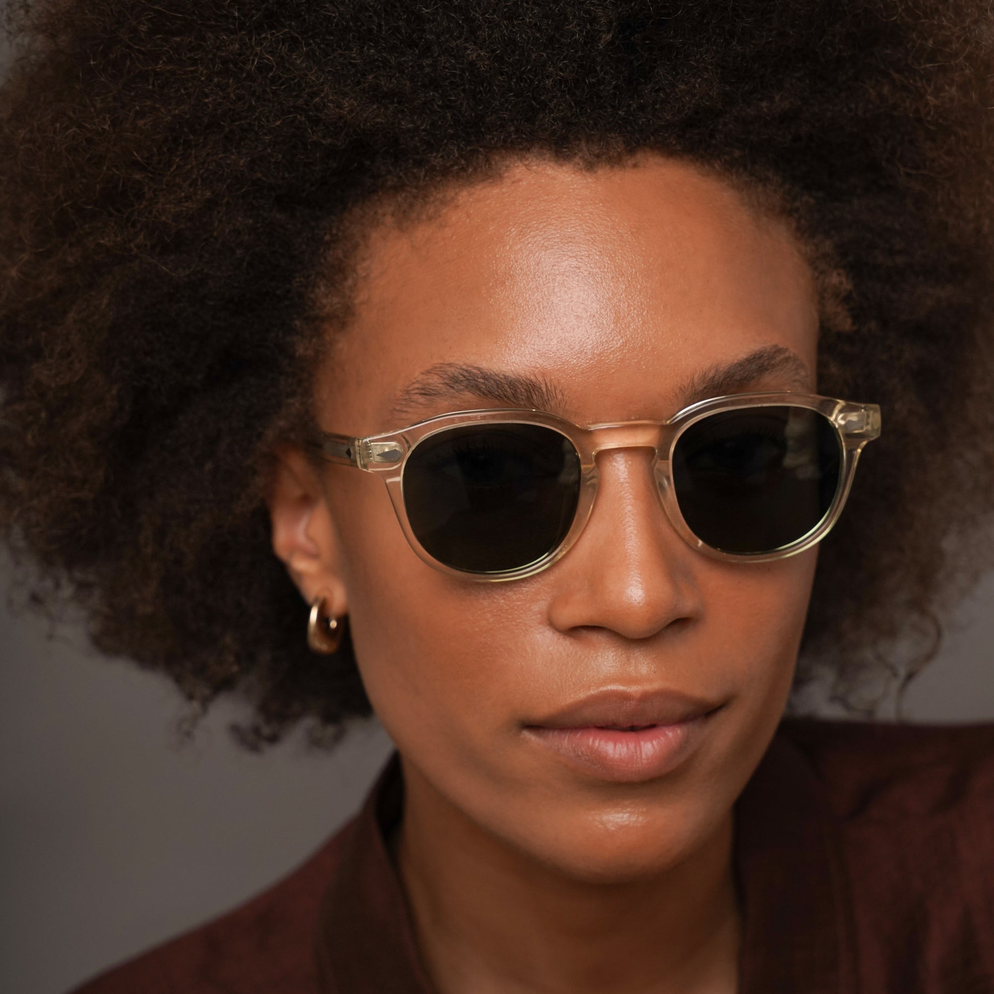 A close-up portrait of a person with dark curly hair wearing round sunglasses with a gold-tone metal frame and dark lenses, against a plain gray background.