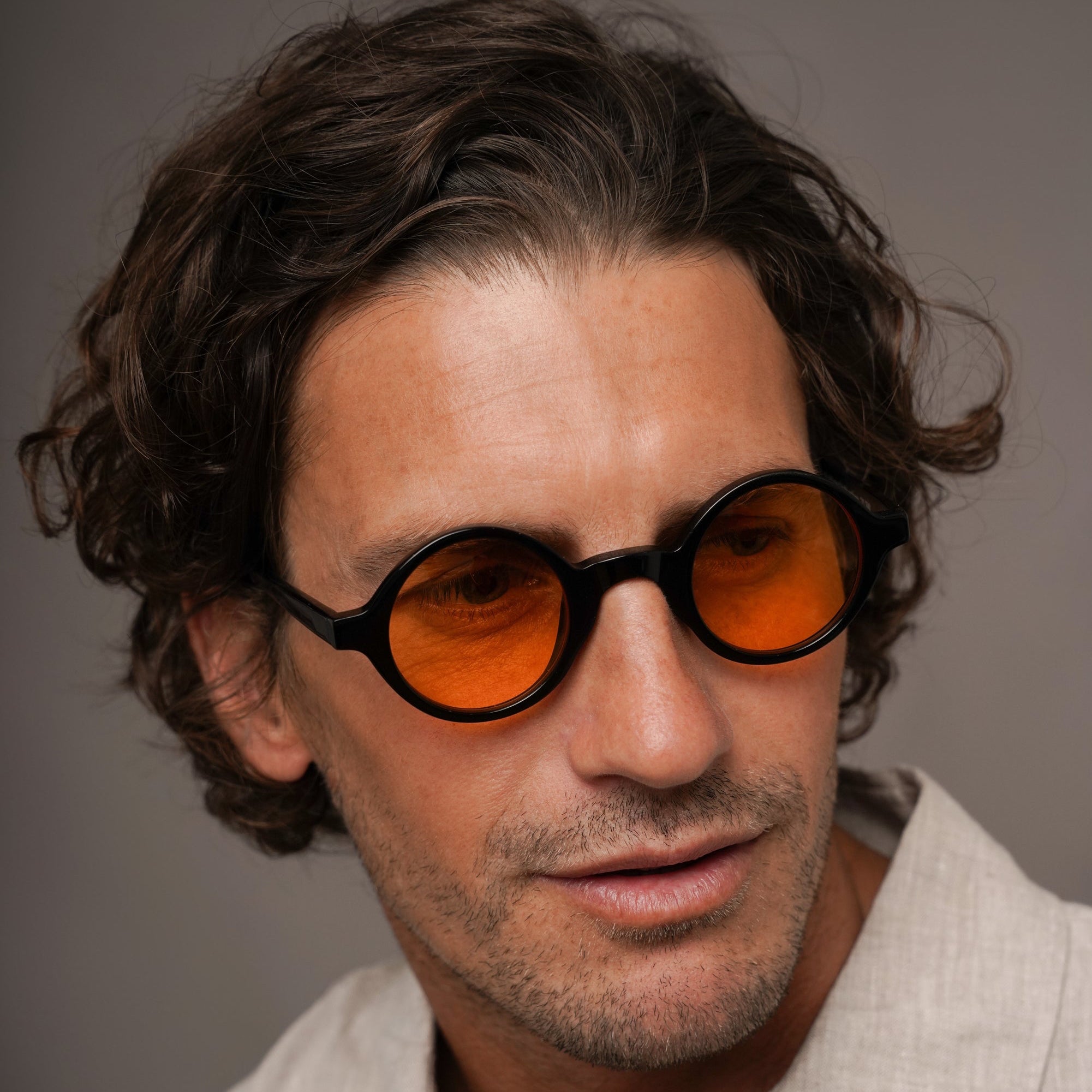 A close-up portrait of a man with dark curly hair wearing round black-framed sunglasses with vibrant orange lenses, set against a neutral gray background.