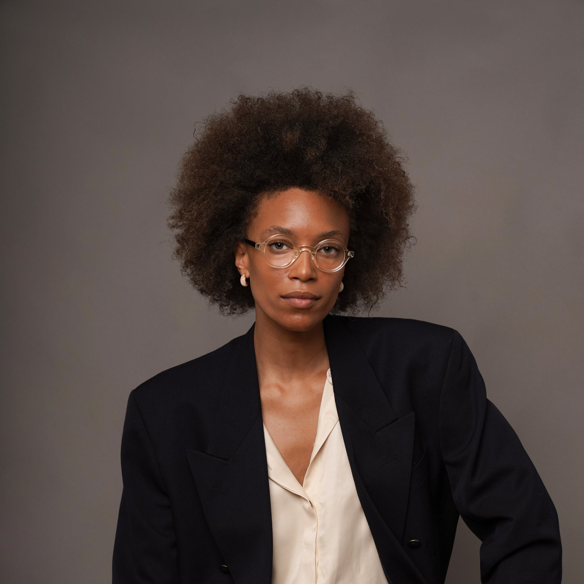A woman with an afro hairstyle and glasses sits on a stool wearing a dark navy blazer, cream blouse, and dark navy trousers against a plain gray studio background.