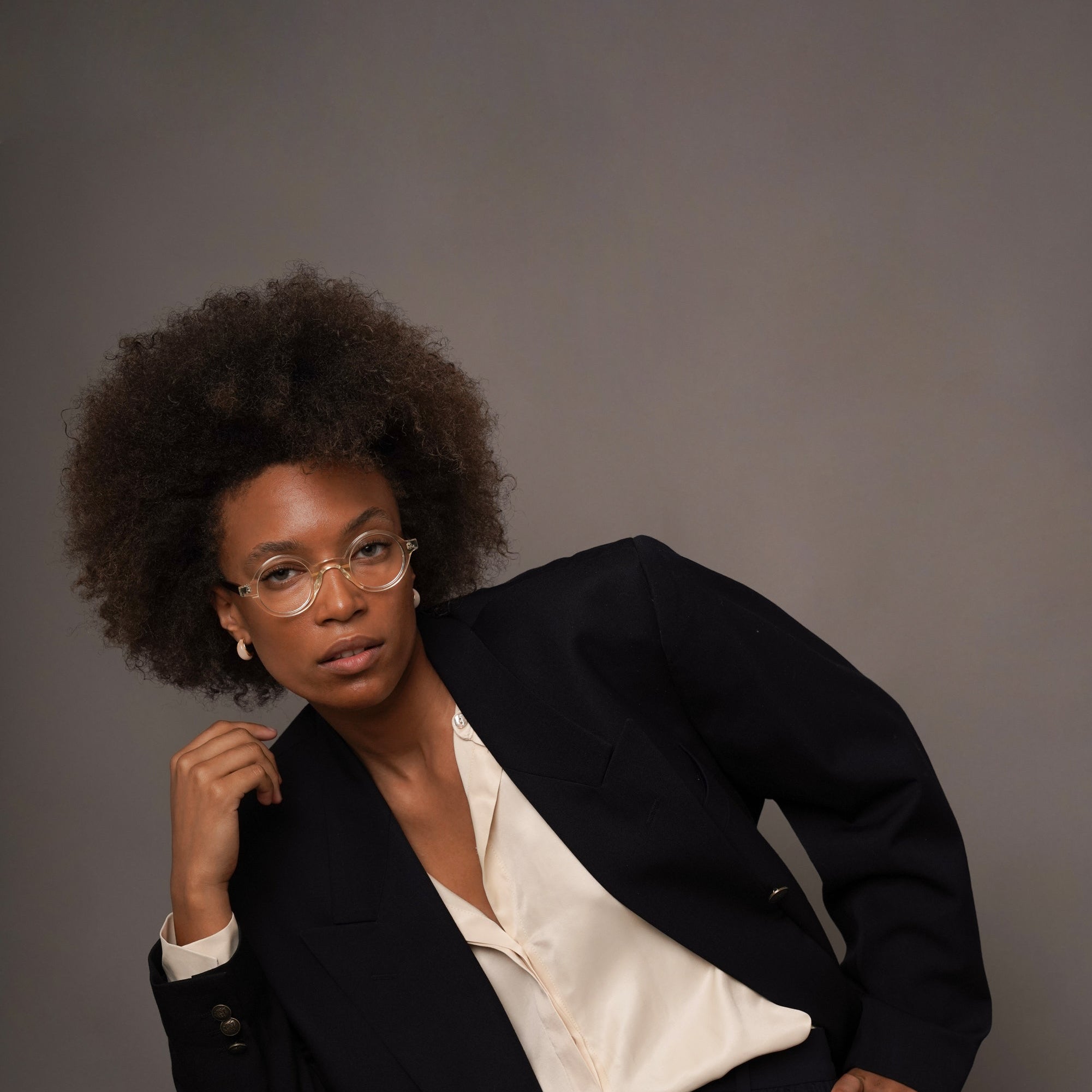 A woman with an afro hairstyle and glasses, wearing a dark navy blazer over a cream satin blouse and matching navy trousers, sitting on a stool against a solid gray studio background.