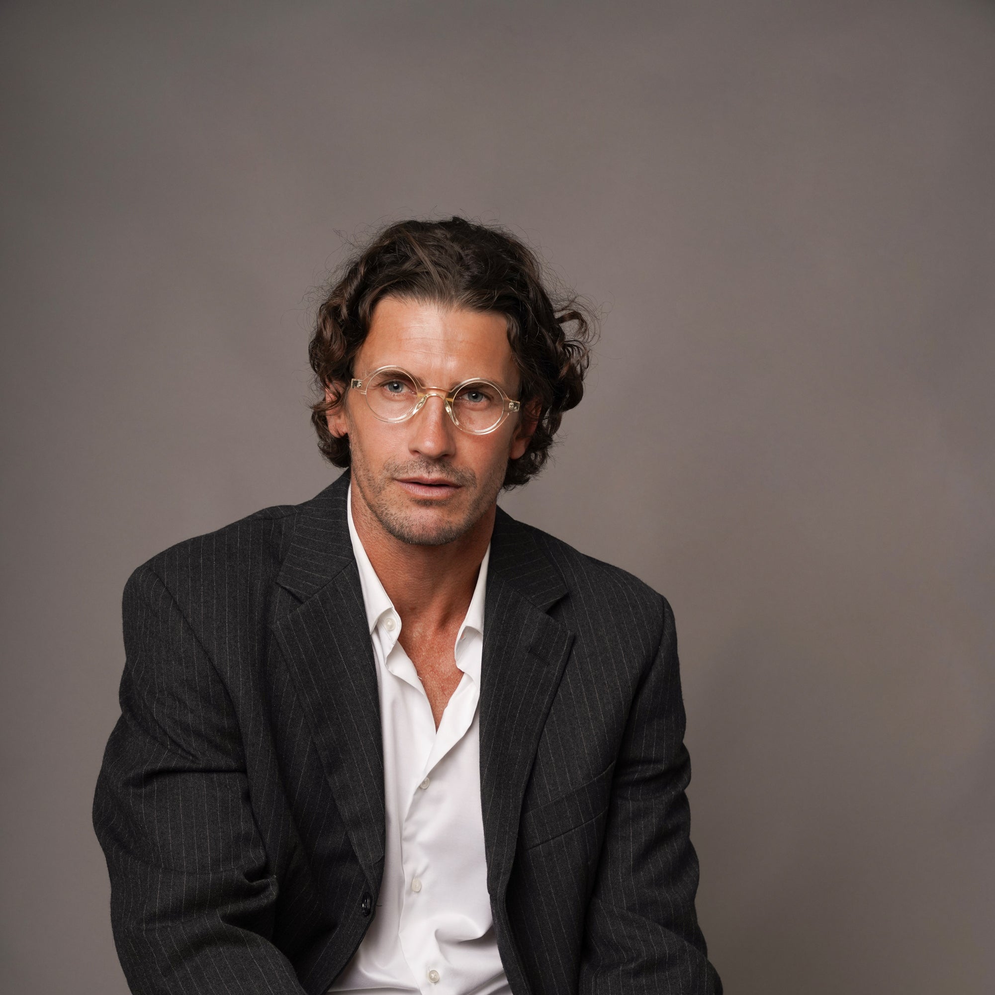 A man wearing a dark pinstripe blazer, white shirt, and dark trousers sits against a plain taupe-gray studio background.